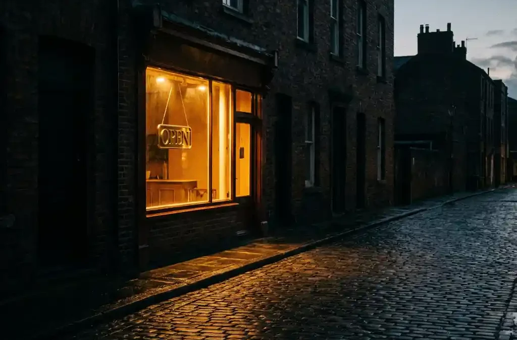 Verlichte winkel op een rustige kasseienstraat, warm gouden licht valt door het raam op grijze stenen, omliggende gebouwen donker.